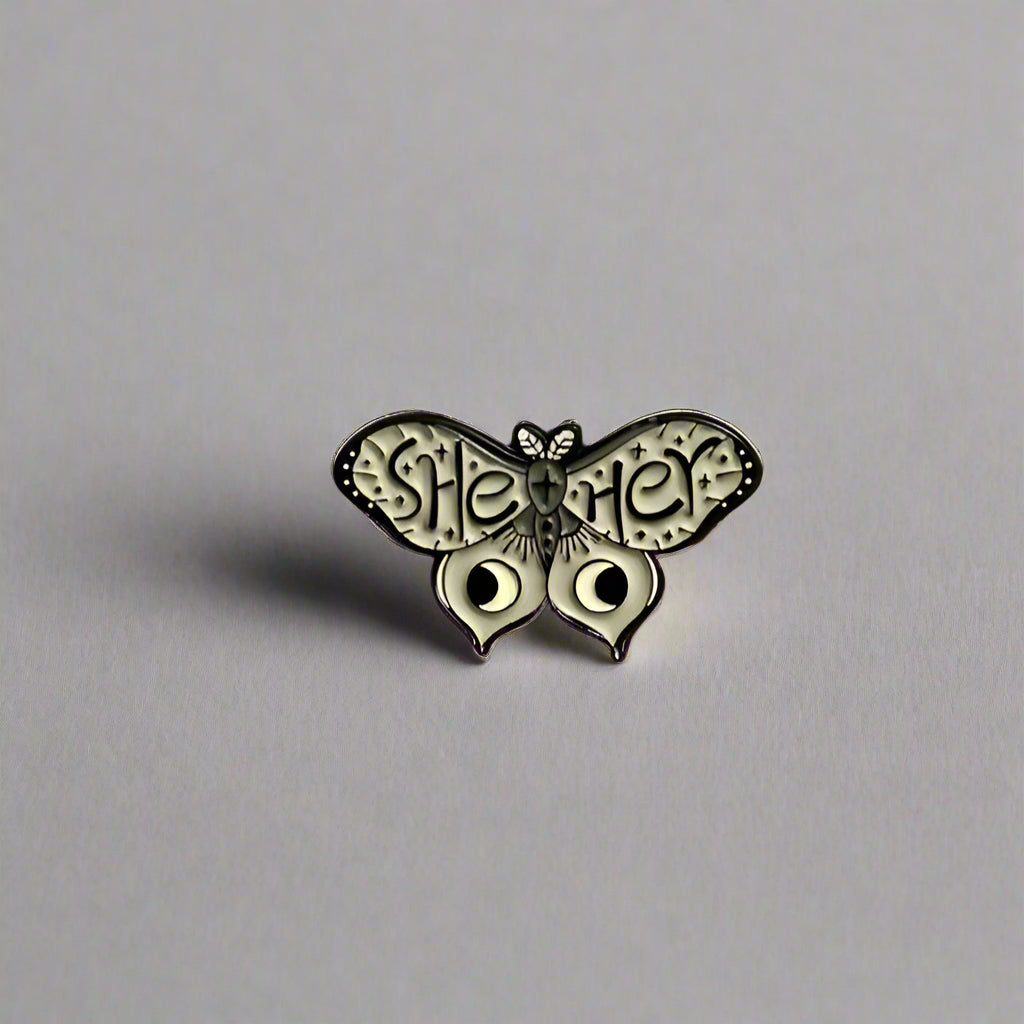Metallic butterfly-shaped pin with 'She/her' text on a beige background