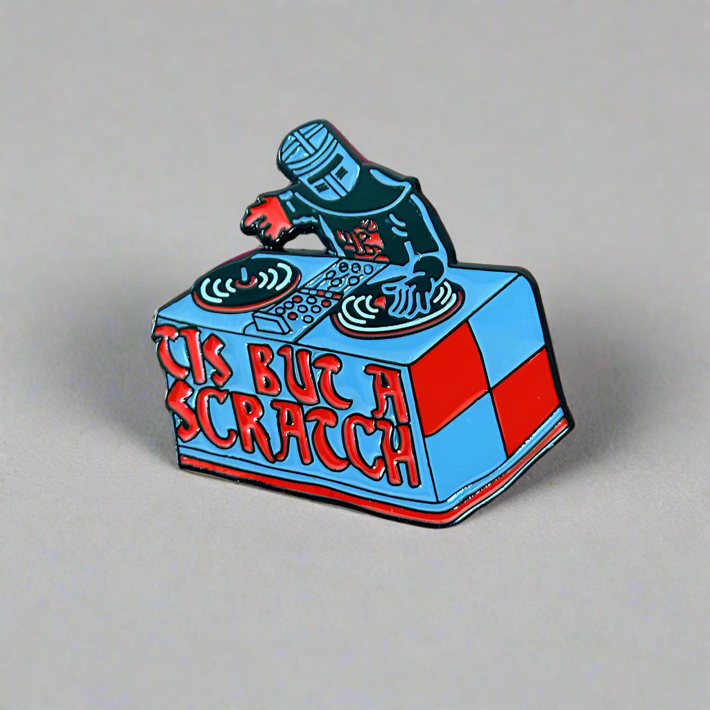 Pin with DJ console design and 'This But A Scratch' text on a wooden surface