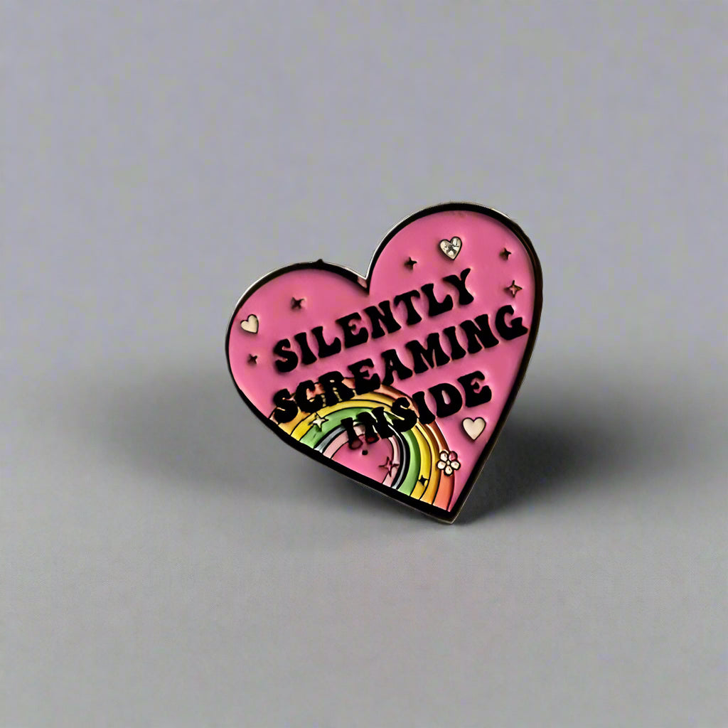 Heart-shaped pink enamel pin with text 'SILENTLY SCREAMING INSIDE' and rainbow design on a beige background