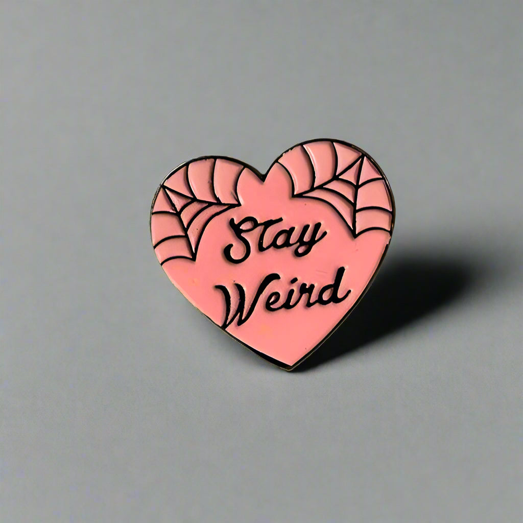 Heart-shaped pink enamel pin with 'Stay Weird' text on a gray background