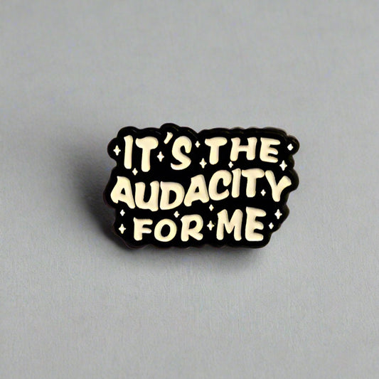 Black and white enamel pin with 'IT'S THE AUDACITY FOR ME' text on a gray background