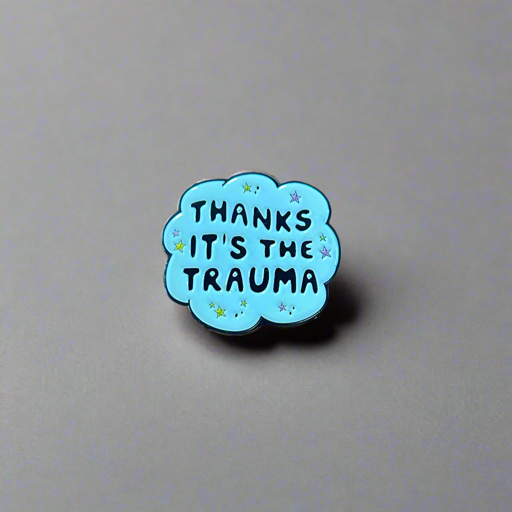 Blue cloud-shaped pin with 'THANKS IT'S THE TRAUMA' text on a wooden surface